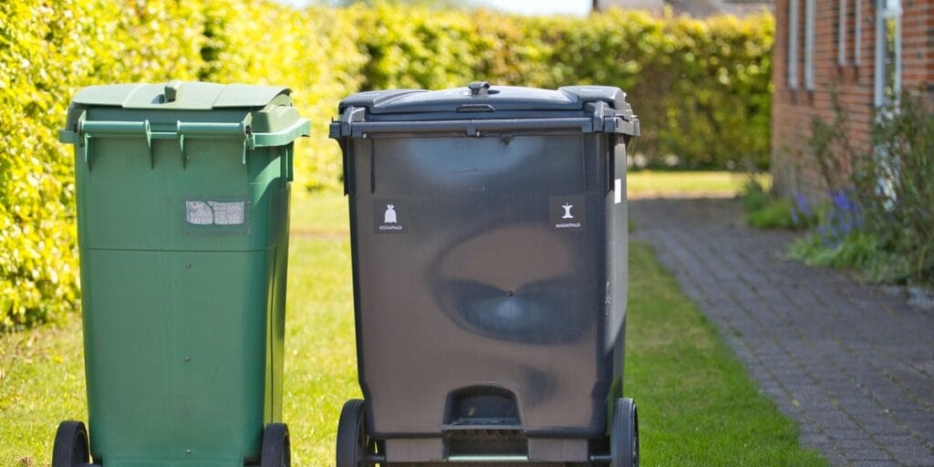 Two closed wheeled trash bins, one green and one dark gray, sit on a lawn beside a paved walkway with a hedge and house in the background.