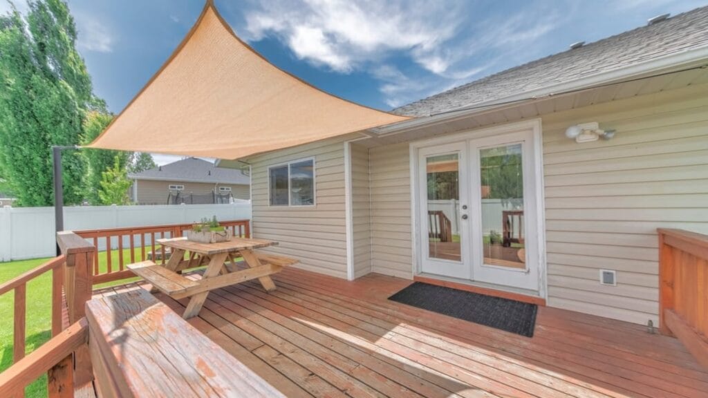 A wooden backyard deck with a triangular shade sail covering a picnic table, next to a house with white French doors, a black door mat, and a lawn beyond the railing.
