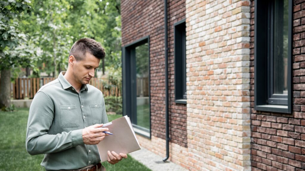 A man in a green shirt stands beside a brick home, reading through papers on a folder while checking the property.