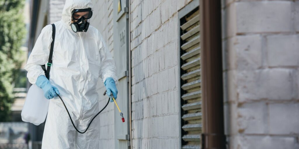 A technician in a full protective suit and respirator sprays chemical along the base of a building wall.