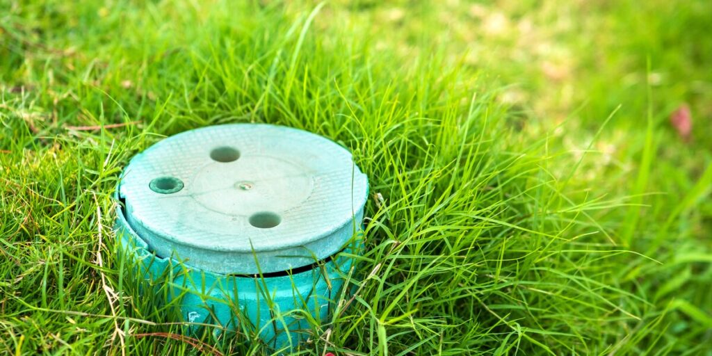 A round teal monitoring station lid sits low in a patch of thick green lawn grass.