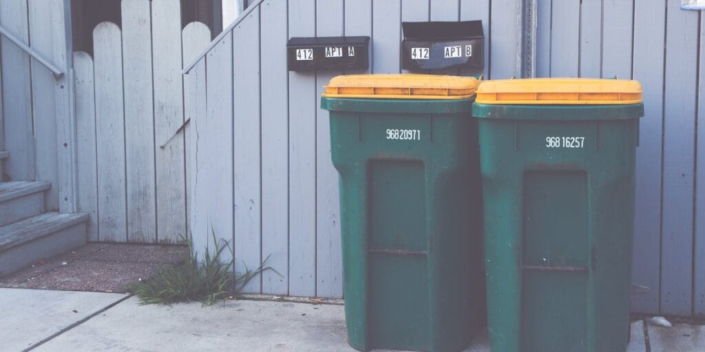 Two green trash bins with yellow lids sit against a wooden fence by a home's back door.