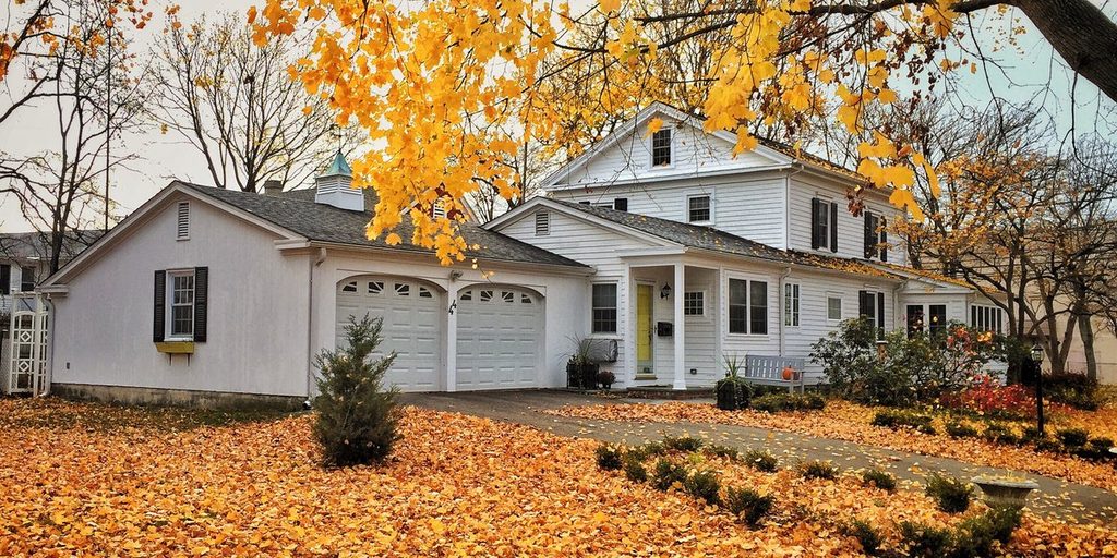 A white two story house with an attached garage sits behind a front yard completely covered in fallen orange and yellow leaves, with bare and golden trees overhead.