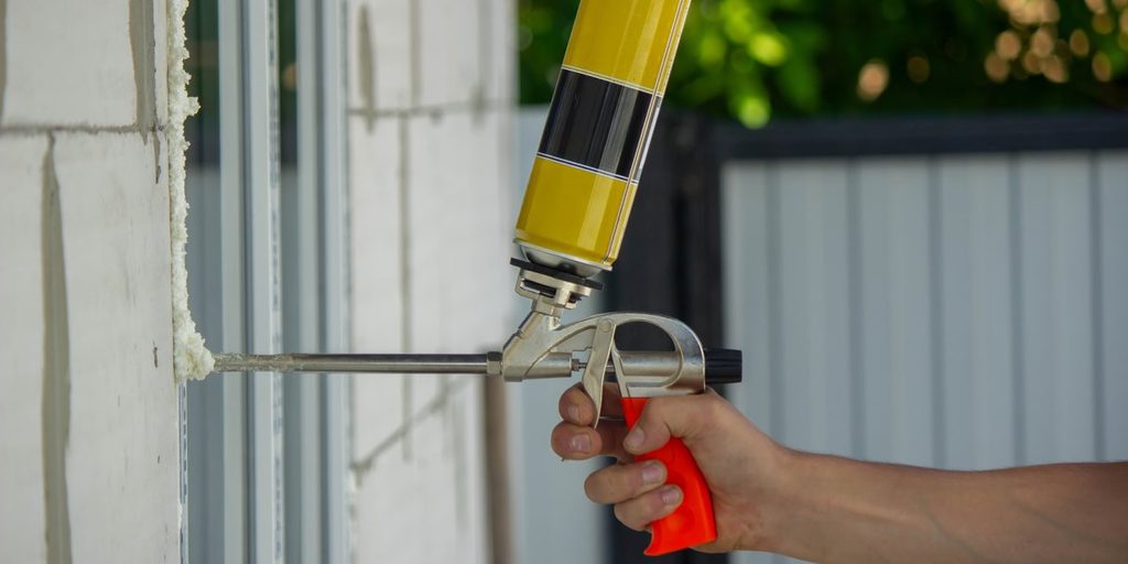 A hand holds a foam gun and injects white expanding foam sealant into a gap beside an exterior door frame.
