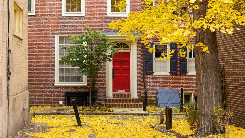 A brick rowhouse with a bright red front door and dark shutters, its front yard buried under a thick layer of fallen yellow leaves.