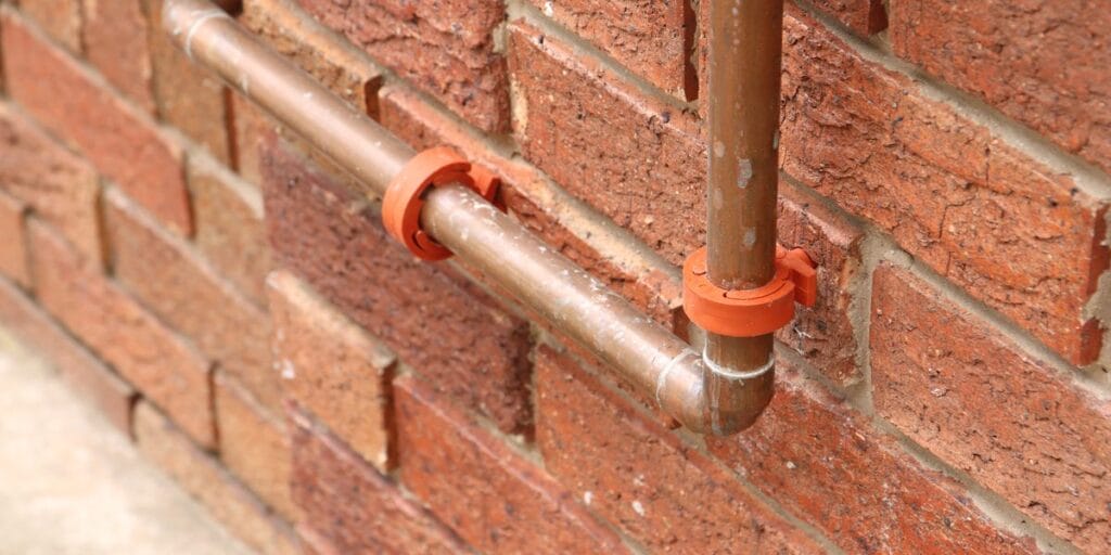 Copper pipes with orange clips run across a red brick wall and bend downward near the bottom.