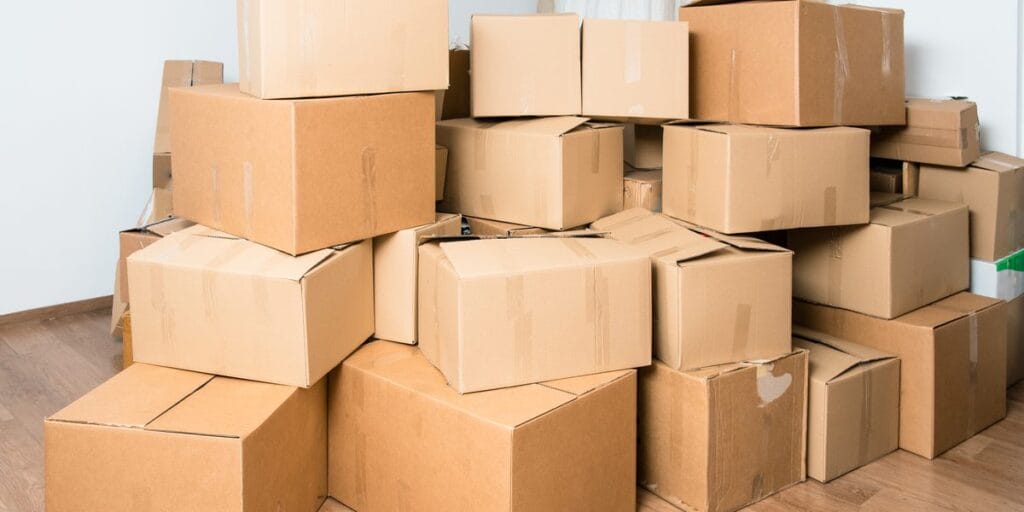 A tall, messy stack of closed cardboard boxes sits against a white wall on a wood floor.