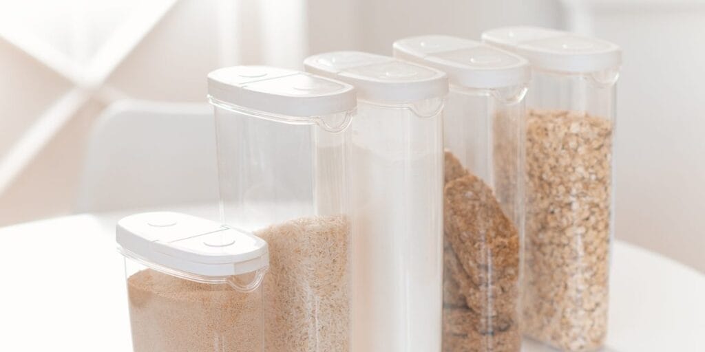Five tall clear containers with white lids hold rice, flour, oats and other dry goods on a bright white counter.